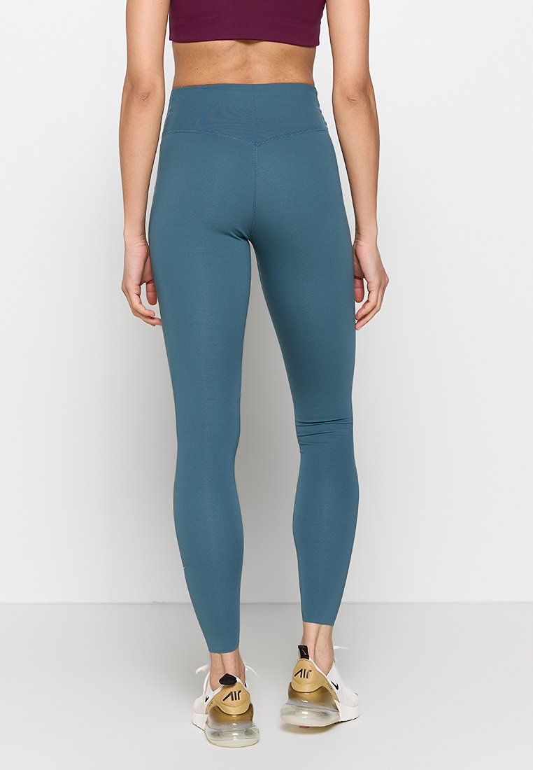 Mujer con leggings de talle alto en color teal y zapatillas blancas está de pie frente a una pared blanca con las manos relajadas a los lados.