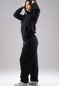 Woman adjusting hood on black zip-up hoodie, wearing matching black sweatpants and silver athletic shoes, standing against plain background.