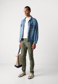 Veste en denim, t-shirt blanc, pantalon vert olive, portant un sac fourre-tout beige avec des accents bleu marine, et chaussé de baskets beiges à rayures.