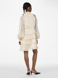 Vrouw in een crèmekleurige gelaagde chiffon jurk met lange mouwen, staande in zwarte hoge hakken, gezien vanaf de achterkant.