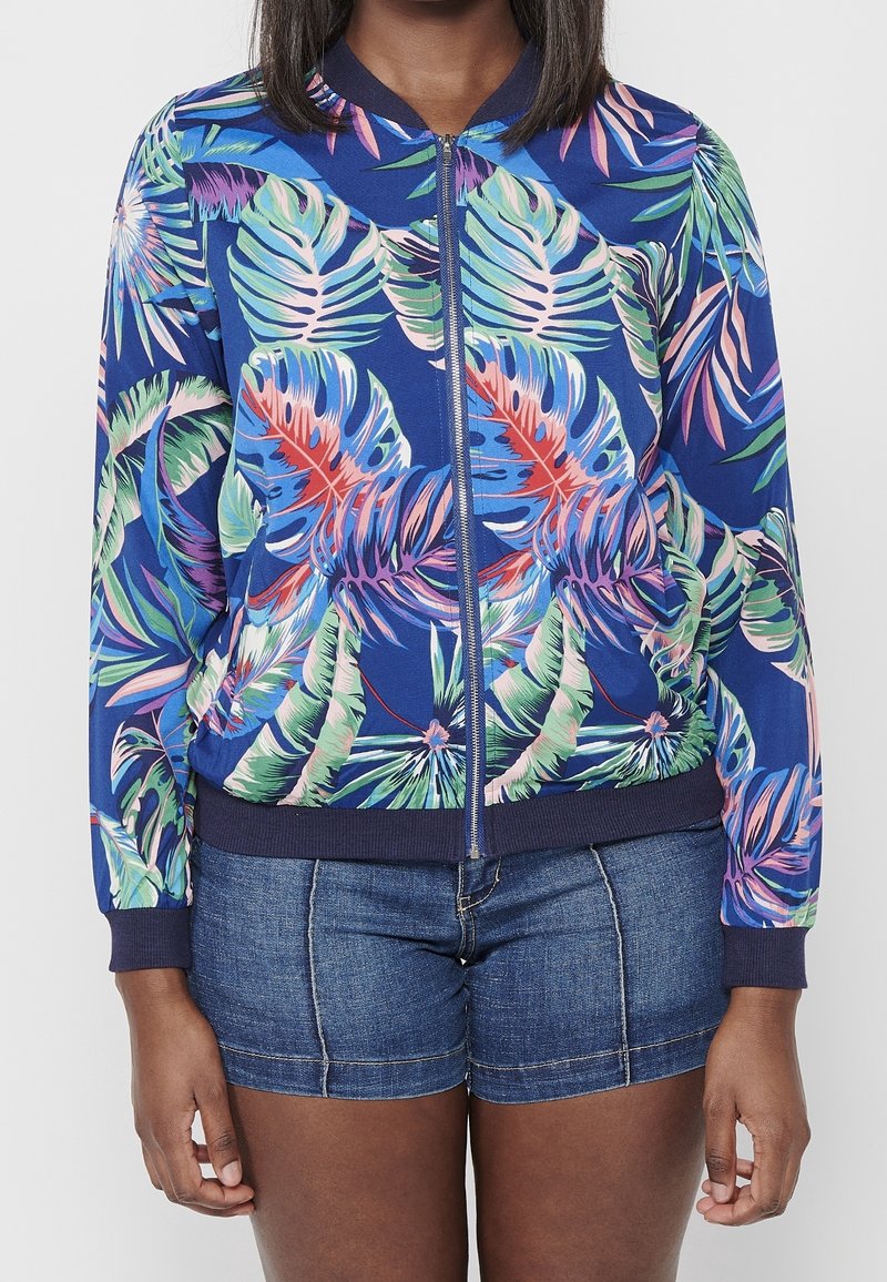 Femme portant une veste bleue zippée avec des motifs de feuilles tropicales multicolores et un short en jean, debout devant un fond uni.