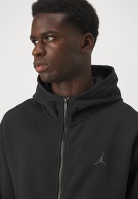 Sort zip-up hættetrøje med høj krave, lavet af struktureret stof, med sølvfarvet lynlås og en diskret grå Jordan-logo på brystet.