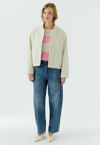 Frau trägt eine cremefarbene Jacke über einem rosa-beigefarben gestreiften Pullover, blaue Jeans und beige spitze Schuhe und steht vor einem einfarbigen Hintergrund.