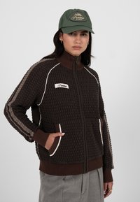 Brun teksturert hettejakke med glidelås, ribbestrikkede mansjetter og krage. Har beige detaljer og to sidelommer. Følges med en grønn caps.