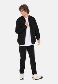 Czarna kurtka bomberka z okrągłym dekoltem, noszona na białej koszulce. Do tego czarne dżinsy i beżowe sneakersy z zielonymi akcentami.