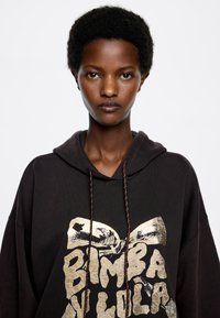 Sudadera negra de gran tamaño con un estampado gráfico dorado, que presenta un cordón con cuentas de colores. Tejido suave con un corte holgado.
