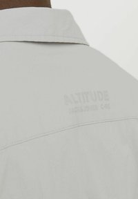 Helle graue Jacke mit glatter Textur, ausgestattet mit einem spitzen Kragen und erhabenem Logotext auf dem Rücken. Doppelt genähte Nähte sind sichtbar.