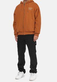 Sudadera naranja con cremallera y bolsillo canguro, combinada con pantalones cargo negros con una rodilla desgastada y zapatillas blancas.