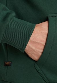 Green sweatshirt featuring ribbed cuffs and a front pocket, made from soft fabric with a tag detail located near the pocket.