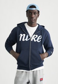 Μπλε σκούρο φούτερ με φερμουάρ και λευκό λογότυπο "nike" στο στήθος, σε συνδυασμό με λευκό t-shirt και γκρι παντελόνι φόρμας με κόκκινο έμπλαστρο Nike. Ανοιχτό μπλε καπέλο φορεμένο ανάποδα.