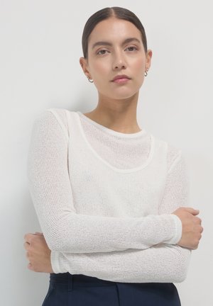 Top bianco a maniche lunghe, leggero, in maglia con scollatura a barchetta e tessuto testurizzato, indossato con pantaloni blu scuro, braccia incrociate.