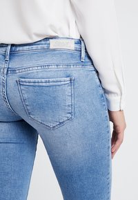 Pantalones vaqueros de denim azul claro con corte slim; cuentan con bolsillos traseros, una textura desgastada y una etiqueta en la cinturilla.