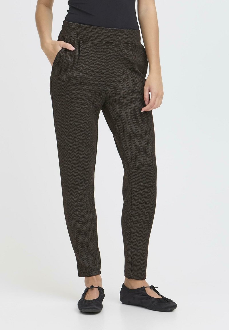 Femme portant un pantalon fuselé à motif chevron marron foncé et des ballerines noires, debout sur un fond blanc.