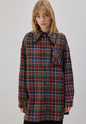 Jeune femme aux cheveux blonds portant une chemise large à carreaux colorés avec un grand col et une poche avant, debout devant un fond uni.