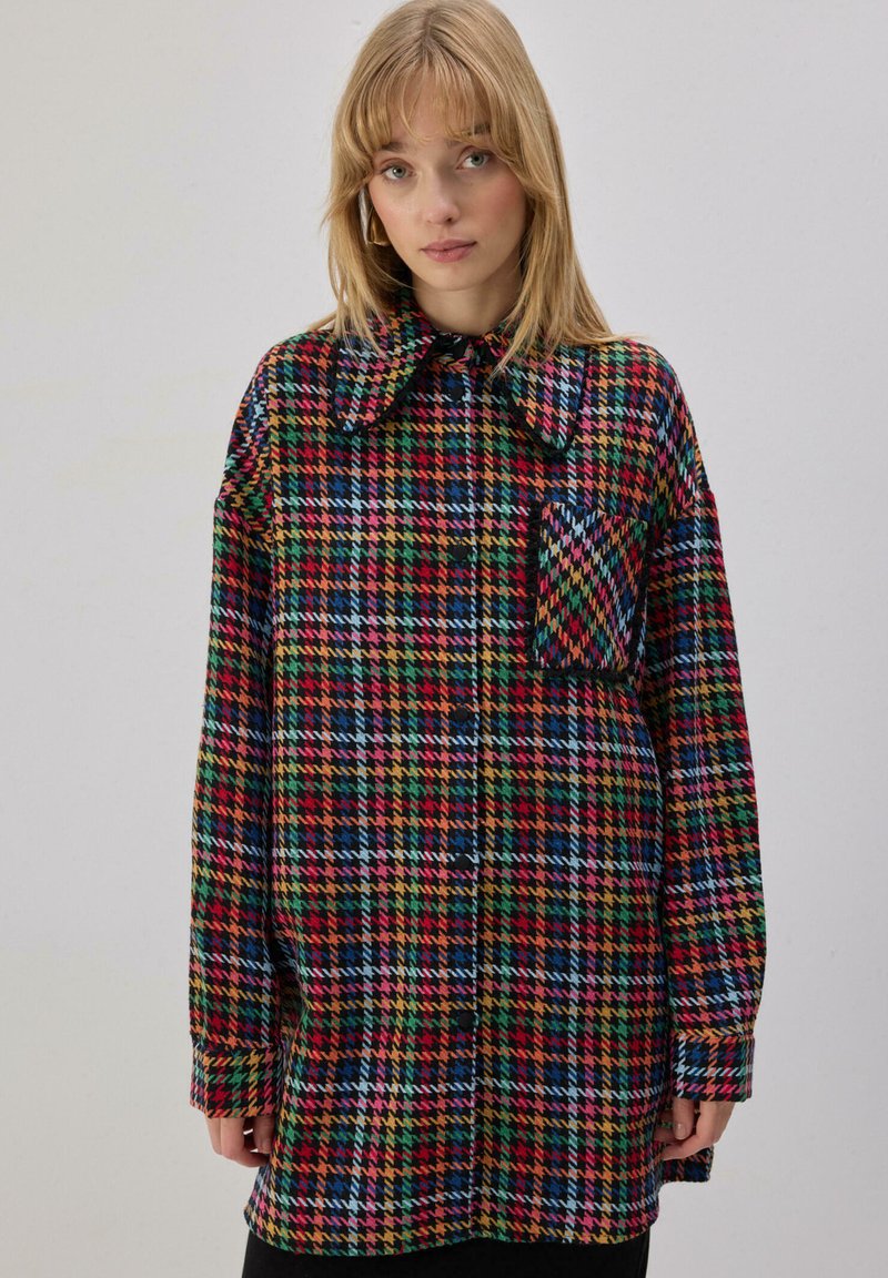 Νεαρή γυναίκα με ξανθά μαλλιά, φορούσα πολύχρωμο υπερμεγέθες πουκάμισο με καρό σχέδιο, μεγάλο γιακά και μπροστινή τσέπη, στέκεται μπροστά σε απλό φόντο.