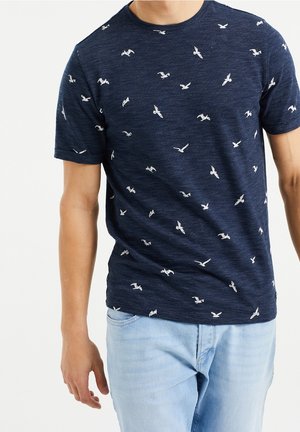 Granatowa koszula z krótkim rękawem o teksturowanej tkaninie z białymi wzorami ptaków. Noszona z jasnoniebieskimi dżinsami.