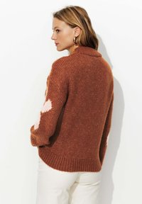Pull en tricot de couleur rouille avec un col haut, présentant des motifs de cœurs roses sur les manches et une coupe décontractée avec un ourlet côtelé.