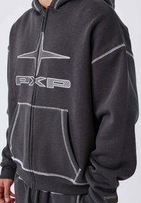 Sudadera gris con cremallera, costuras blancas, bolsillos frontales y un logo en el pecho. Cuenta con capucha y puños acanalados para un mejor ajuste.