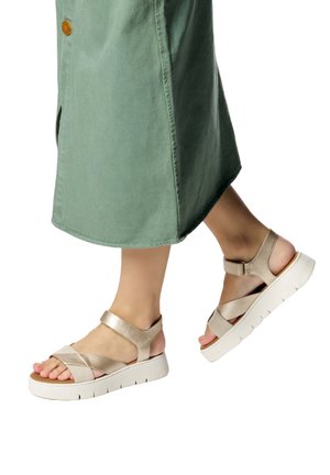 Piedi che indossano sandali con zeppa beige e cinturini in Velcro, abbinati a una gonna midi verde, su sfondo bianco.