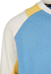 Gros plan d’un sweat-shirt bicolore avec devant bleu, manches crème et accents jaunes près de l’épaule et du côté.