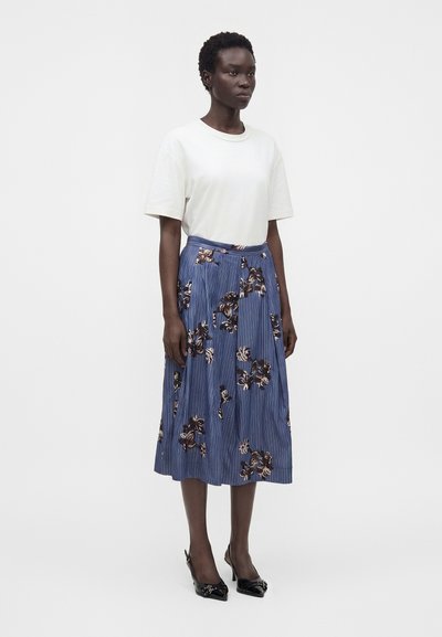 Femme portant une chemise blanche à manches courtes, une jupe midi bleue à motifs floraux et des talons noirs à bout pointu, debout devant un fond uni.