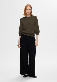 Maglione a maniche lunghe col girocollo di colore verde oliva, con spalle arricciate, abbinato a pantaloni neri a gamba larga e scarpe nere basse.