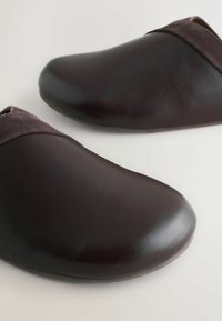 Zuecos de cuero marrón oscuro con un acabado liso, punta redondeada y suaves detalles de ante en la parte superior, con un diseño minimalista.
