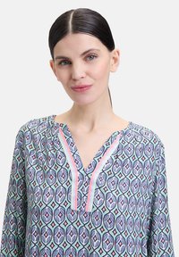 Mehrfarbige gemusterte Bluse mit V-Ausschnitt und dezenten Falten. Enthält geometrische Formen in Blau, Grün und Pink auf einem hellen Hintergrund.