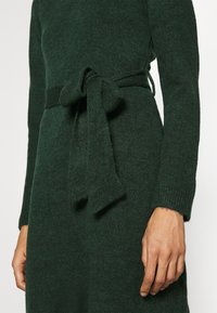 Close-up of dark green knit dress with long sleeves and tied fabric belt at waist, showing part of the wearer's hands.