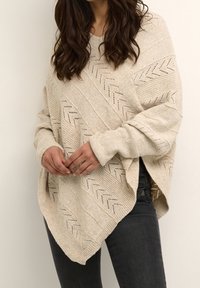 Beige breiponcho met een gestructureerd patroon, met diagonale lijnen en een subtiel chevronontwerp. Ruime pasvorm met zijsplitten en brede mouwen.