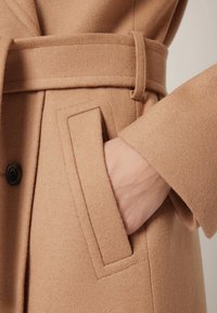 Płaszcz z wełny wielbłądziej z szerokim paskiem, kieszeniami na klapki i zapięciem na guziki. Miękka tekstura, dopasowany krój i czyste linie.