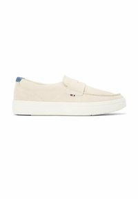 Mocasín beige sin cordones con suela gruesa de goma blanca, lengüeta azul en el talón y pequeña etiqueta de tela con rayas rojas, blancas y azules en el lateral.