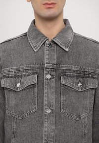Gray denim jacket featuring a button-up front, two chest pockets, and a pointed collar. Silver hardware and textured fabric detail are visible.