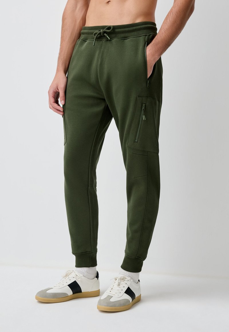 Uomo che indossa pantaloni da jogging verde scuro con tasche laterali con cerniera e sneaker bianche, in piedi davanti a uno sfondo semplice.