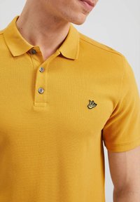 Gul bomullspolo skjorta med en texturerad väv, tre-knapplås och en liten svart broderad logotyp på vänster bröst.