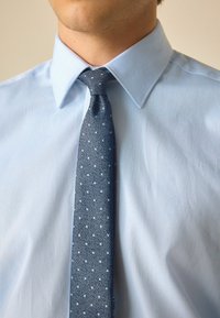 Hellblaues Hemd aus strukturiertem Stoff, mit einer schmalen, dunkelblauen Krawatte mit kleinen hellblauen Punkten.