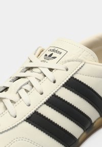 Λευκό δερμάτινο αθλητικό παπούτσι με μαύρο σχέδιο τριών γραμμών, εμφανές λογότυπο Adidas στη γλώσσα, υφασμένα διακοσμητικά στοιχεία και μπεζ σόλα.