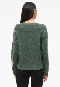 Pull en tricot vert avec un motif en losanges, col rond et ourlet côtelé. Manches longues avec une finition texturée. Jean noir visible à la taille.