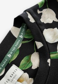 Czarna bluzka w kwiaty z kremowymi kwiatami i zielonymi liśćmi. Miękka tkanina z wplecioną metką, na której widnieje napis "Ted Baker London". Teksturowany dekolt.
