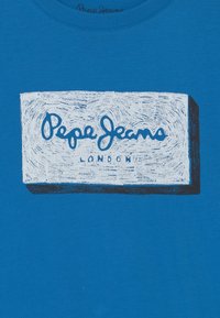 Blå bomulls t-shirt med en vit rektangulär grafik med "Pepe Jeans" och "London" i stiliserad text. Slät textur.