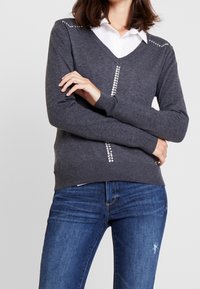 Grå stickad tröja med V-ringning, prydd med strass på axlar och framstycke. Kombinerad med blå jeans, visar upp en figurnära silhuett.