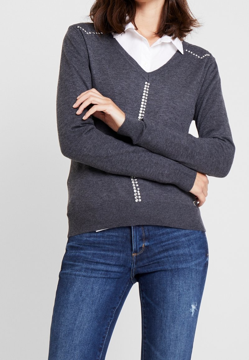 Grå stickad tröja med V-ringning, prydd med strass på axlar och framstycke. Kombinerad med blå jeans, visar upp en figurnära silhuett.