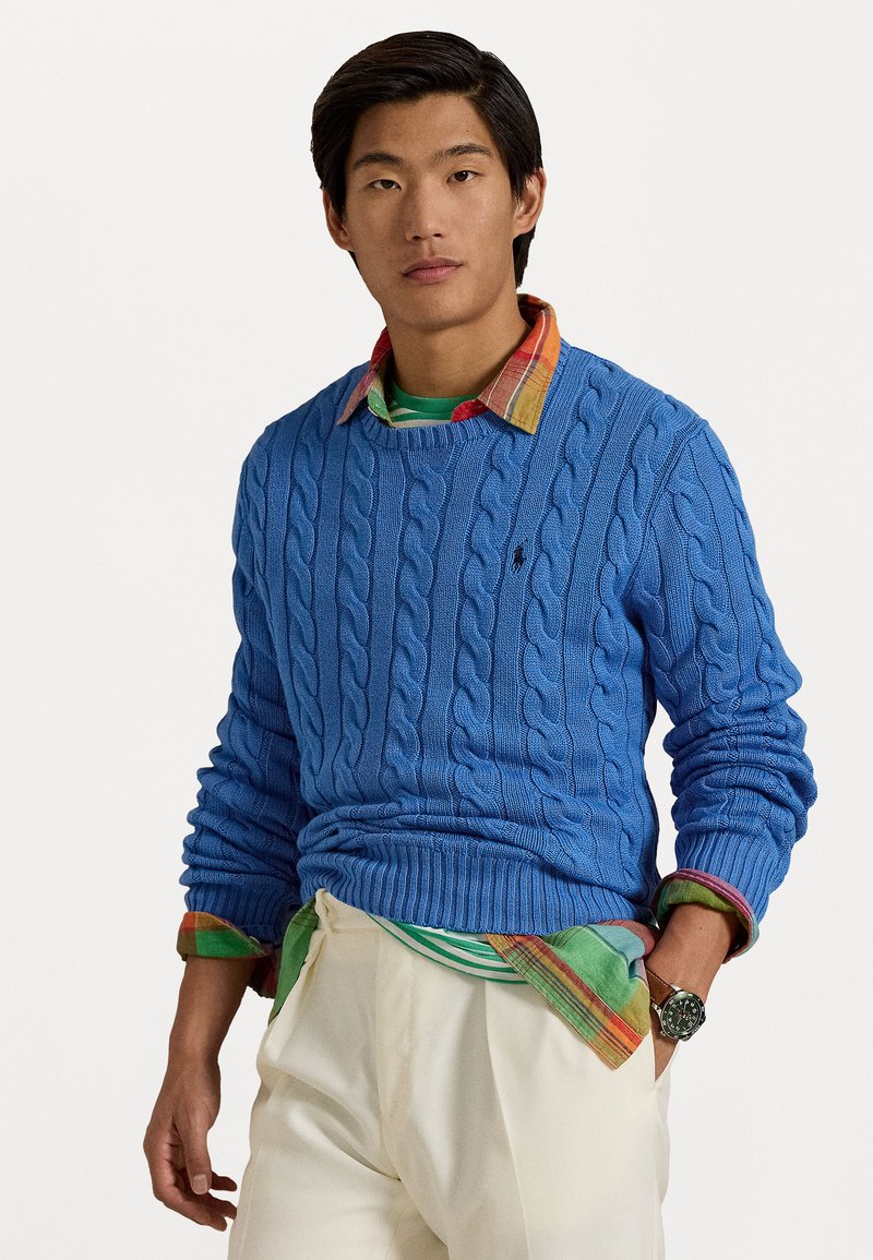 Cable-knit blue sweater with a round neckline, worn over a multicolored plaid shirt. The outfit features cream-colored trousers and a watch.