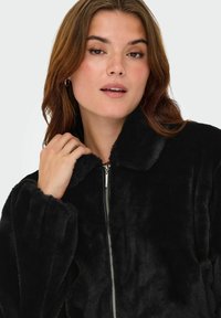 Zwarte faux fur jas met een ritssluiting, hoge kraag en ruime mouwen. Zachte textuur met een subtiele glans, ideaal voor koel weer.