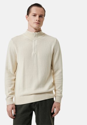 Young man with slicked-back hair wearing a cream-colored, long-sleeve half-zip sweater and dark pants, standing against a plain background.