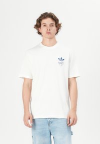 T-shirt en coton blanc à manches courtes, présentant un design de logo bleu. La coupe est décontractée et la texture semble lisse.