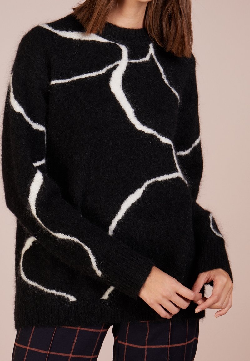 Pull en maille noire avec motif abstrait blanc, col rond et poignets côtelés. Texture douce avec une coupe ample.