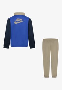 Nike trenirka, ki vsebuje modro jakno s temnonavdnimi rokavi in bež hlačami. Jakna ima izrazit logotip na hrbtni strani in elastične manšete.