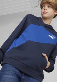 Granatowa bluza z kapturem z niebieskim akcentem, wyposażona w kieszeń kangurką oraz żebrowany dół. Logo PUMA w białym kolorze umieszczone na piersi.