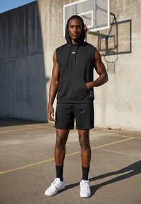 Persona atlética con sudadera sin mangas negra y pantalones cortos, zapatillas blancas, de pie en una cancha de baloncesto al aire libre cerca del aro y una pared de concreto.